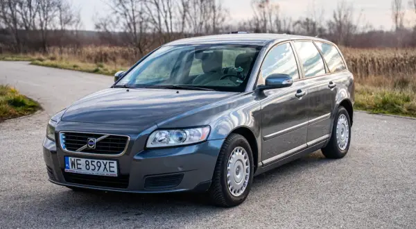 Szary v50 volvo na drodze, otoczony suchą trawą i drzewami.