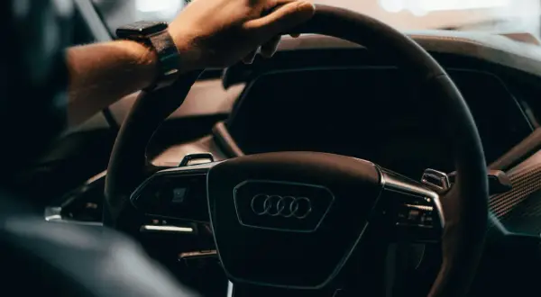 Dłoń na kierownicy Audi, z zegarkiem na nadgarstku. W tle widać fragment deski rozdzielczej, sugerujący wnętrze nowoczesnego samochodu, jak w tabeli z cenami rynkowymi samochodów używanych.