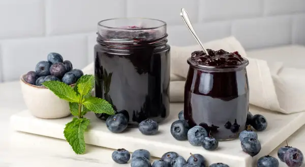 Due barattoli di marmellata di mirtilli, uno aperto con un cucchiaino, circondati da mirtilli freschi e foglie di menta.