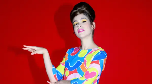 Cosima Ruiz de la Prada, con un peinado voluminoso y maquillaje vibrante, posa con un vestido de estampado psicodélico sobre fondo rojo.