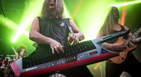 Muzyk gra na keyboardzie na pasku, podczas gdy gitarzysta i perkusista tworzą rockową atmosferę.