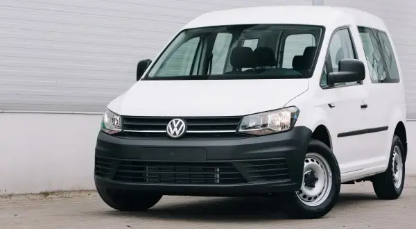 Biały Volkswagen Caddy, samochód osobowy zarejestrowany jako ciężarowy bez kratki, stoi przed szarą ścianą.