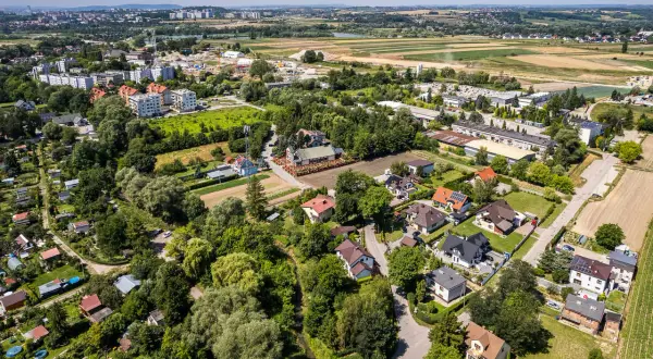 Osiedle domów jednorodzinnych na wzgórzach, otoczone zielenią i polami. W oddali widać zabudowania miejskie.