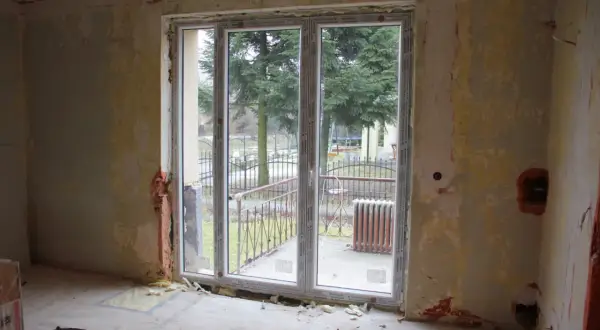 Niedokończona ściana z oknem balkonowym, widok na drzewa i ogrodzenie.