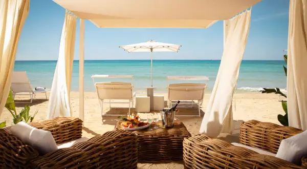 Lussuosa area lounge con divani in vimini, tavolino con frutta e champagne, lettini e ombrellone su una spiaggia di sabbia dorata. Un'oasi tra i migliori beach clubs Puglia.