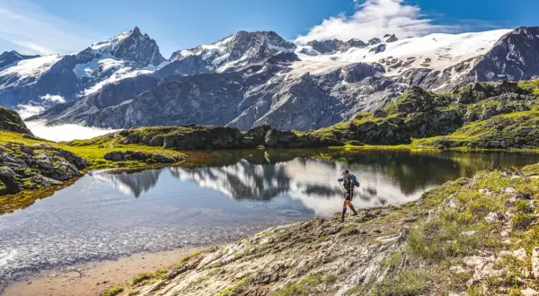 Un randonneur traverse le plateau d'Emparis, un lac alpin reflétant des montagnes enneigées sous un ciel bleu.