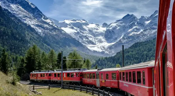 Röda tåg slingrar sig genom en grönskande dal med snöklädda berg i bakgrunden. En drömresa med nattåg genom Europa.