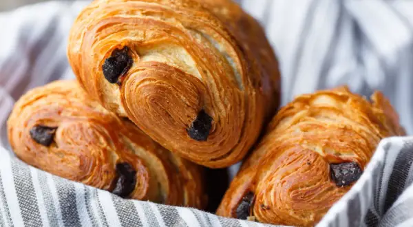 Tre deliziosi pain au chocolat dorati, con strati sfogliati e gocce di cioccolato fondente, adagiati su un canovaccio a righe.
