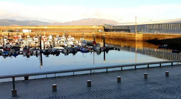 Barcos pesqueros amarrados en el puerto de Finisterre, con un edificio moderno al fondo y montañas en la distancia.
