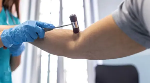 Lekarz w niebieskich rękawiczkach bada odruch TOB u pacjenta, używając młoteczka neurologicznego.