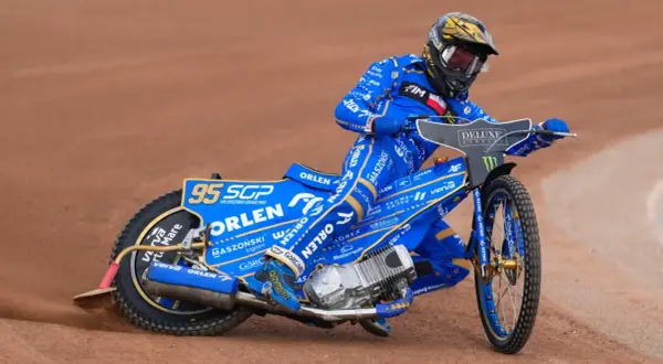Bartosz Zmarzlik w niebieskim kevlarze z numerem 95, na motocyklu Orlen, ściga się na żużlu. Jego wzrost nie przeszkadza w dynamicznej jeździe.
