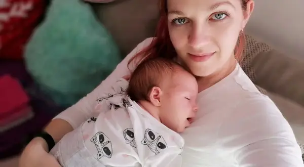 Mama tuli otula swoje śpiące dziecko, czułe kangurowanie dziecka w białym pajacyku z motywem zwierzątek.