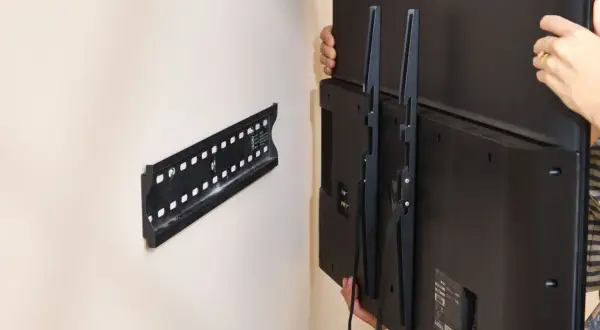 Hands carefully lift a TV, showing how to remove TV from wall mount. The wall bracket is visible.