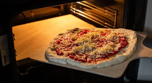 Pizza z sosem i serem na desce, gotowa do włożenia na rozgrzany kamień szamotowy w piekarniku.