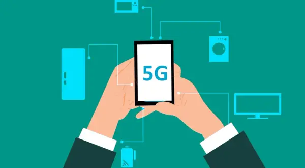 Dłonie trzymają smartfon z napisem 5G, połączony z różnymi urządzeniami domowymi. To pokazuje, co to jest 5G – przyszłość łączności.