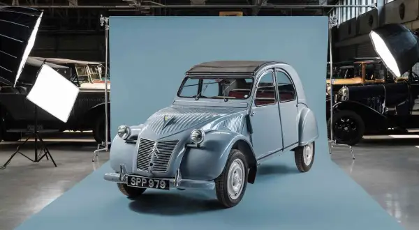 En ljusblå Citroën 2CV står i fokus, redo för fotografering. Bakom den syns andra klassiska bilar och studiobelysning.