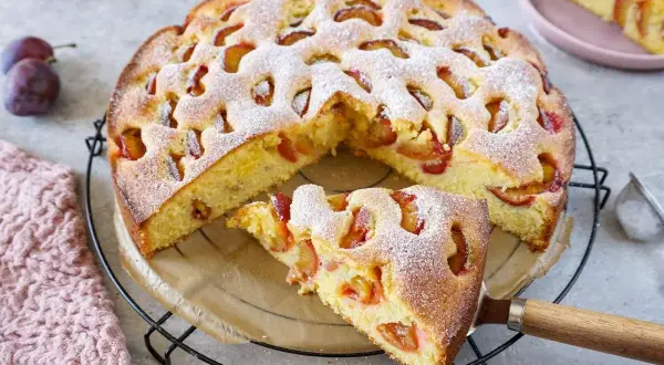 Saftiger pflaumenkuchen mit gefrorenen pflaumen, bestäubt mit Puderzucker. Ein Stück wird gerade herausgenommen.