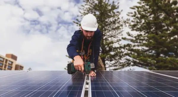 Técnico instala paneles solares, cumpliendo con el real decreto autoconsumo. Edificio y árboles al fondo.