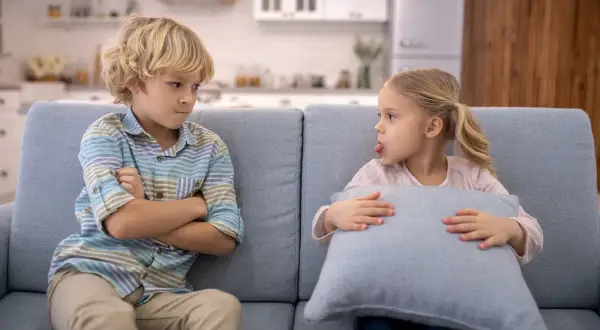 Bambini che litigano sul divano. Lui incrocia le braccia, lei fa la linguaccia con un cuscino.