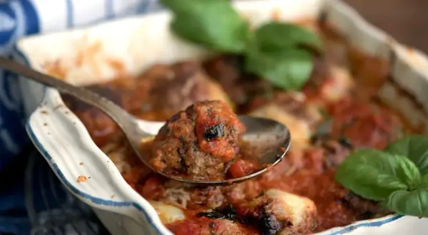 En sked lyfter en köttbulle täckt av mustig tomatsås ur en ugnsform. En härlig pasta med tomatsås väntar!
