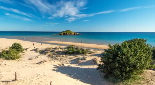 Dune sabbiose e mare turchese, un'isola rocciosa in lontananza. Un assaggio delle spiagge più belle Sardegna, con persone che passeggiano sulla riva.