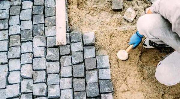 Pracownik w niebieskich rękawiczkach ubija kostkę brukową gumowym młotkiem, tworząc równą nawierzchnię.