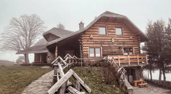 Drewniany dom w stylu góralskim, otoczony mgłą, zaprasza do odpoczynku w Beskidzie Wyspowym. Kamienna ścieżka prowadzi do przytulnego schroniska.