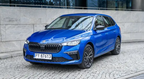 Błękitna Skoda Scala 2024 z czarnymi felgami stoi na brukowanej ulicy przed nowoczesnym budynkiem.
