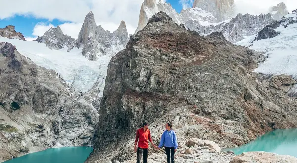 Deux randonneurs se tiennent la main sur un sentier rocheux, admirant un lac turquoise et des montagnes enneigées lors d'une randonnée argentine.