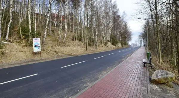 Droga w lesie, z tablicą "Uwaga! Sensacja!", sugerującą miejsce anomalii grawitacyjnej. Po prawej chodnik i ławka.