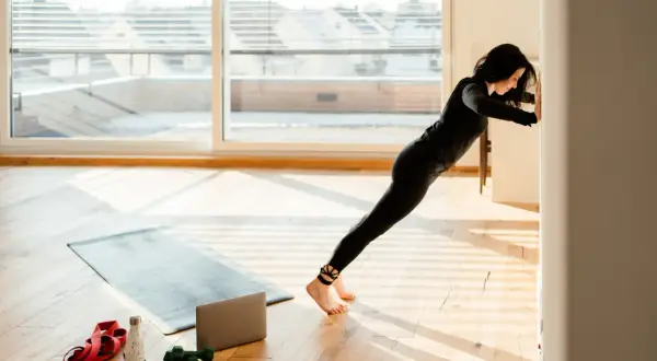 Frau macht Liegestütze an der Wand, um zu lernen, wie trainiert man. Trainingsmatte, Hanteln und Wasserflasche liegen bereit.
