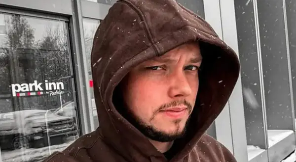 Mężczyzna w brązowej bluzie z kapturem, na tle drzwi hotelu Park Inn. Widać padający śnieg.