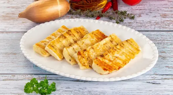 Grillowany ser halloumi na białym talerzu, z dodatkami jak czosnek, cebula, pomidory i chili w tle.