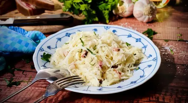 Herzhaftes Krautsalat Rezept mit Speck und frischen Kräutern, serviert auf einem blauen Tellerchen. Dazu Brot, Knoblauch und Petersilie.