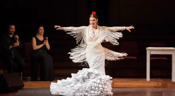 Bailarina de flamenco con vestido blanco y volantes, expresando pasión. A su lado, dos personas aplauden.