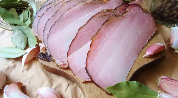 Pokrojony w plastry schab po wędzeniu, otoczony liśćmi laurowymi, czosnkiem i pieprzem.