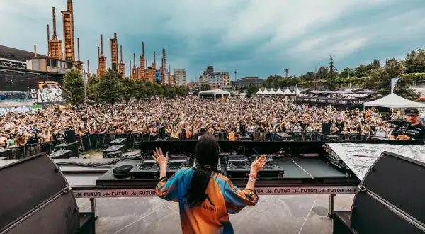 DJ set al futur festival, folla in delirio sotto un cielo nuvoloso, con un'architettura industriale sullo sfondo.