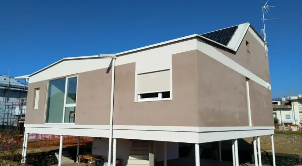 Casa moderna con intonaco argilla, pannelli solari e ampie vetrate. La struttura è rialzata su pilastri bianchi.