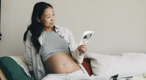 Gravid kvinna tittar på ultraljudsbild, i trimester 2. Hon sitter bekvämt i soffan med sin mage synlig.