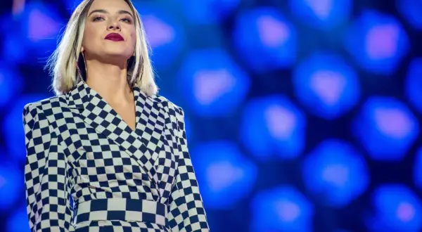 Młoda, amerykańska piosenkarka country w stylowej, szachownicowej marynarce na tle niebieskich świateł.