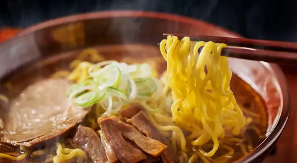 En skål med ångande ramen, med en skillnad på nudlar och pasta som tydligt syns. Nudlarna lyfts med ätpinnar, med kött och grönsaker i buljongen.