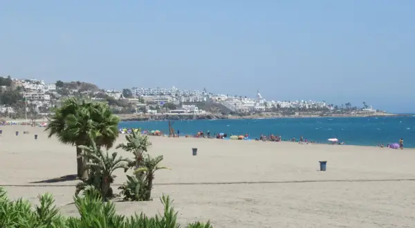 La **playa de la Mena** se extiende con arena dorada y palmeras. Al fondo, un pueblo blanco se asoma al mar azul.