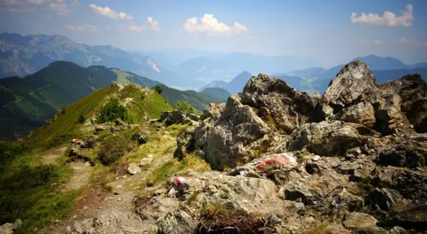 Senderos marcados con rojo y blanco serpentean por las laderas verdes de los Picos de Europa en verano, con rocas imponentes y un valle brumoso al fondo.