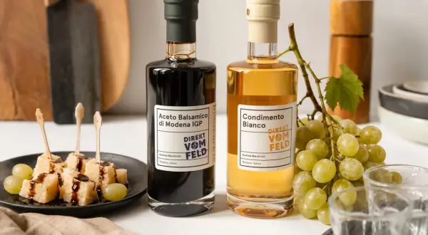 Zwei Flaschen Balsamico und Condimento Bianco, daneben Käsewürfel mit Balsamico und Trauben.