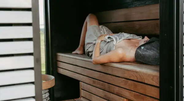 Kobieta w czepku "OF SAUNA" leży na drewnianej ławie w saunie, relaksując się. Dowiedz się, jak oddychać w saunie, by w pełni czerpać z jej dobrodziejstw.