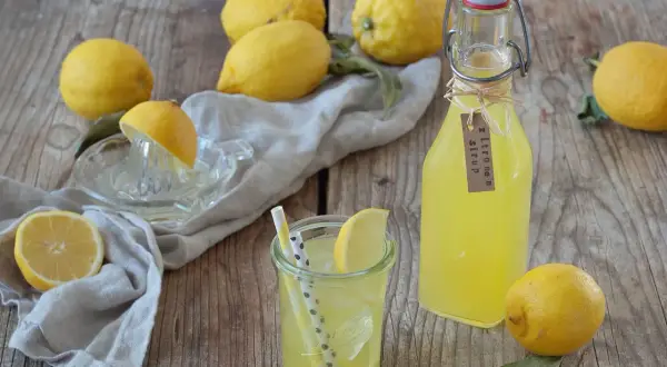 Frische Zitronen, ein Glas mit Eis und Strohhalmen, sowie eine Flasche selbstgemachter Zitronensirup auf Holzhintergrund.
