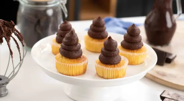 Fem cupcakes med fluffig chokladganache på en vit tårtfat. Ingredienser för chokladganache syns i bakgrunden.