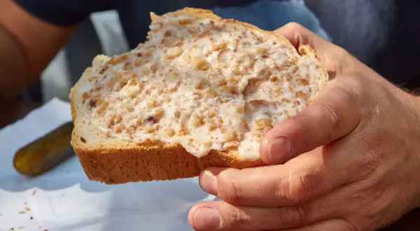 Kromka chleba posmarowana smalcem z kawałkami boczku, trzymana w dłoniach. Obok ogórek kiszony.