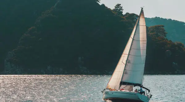 Poppa barca a vela bianca solca acque scintillanti, con una rigogliosa collina verde sullo sfondo.