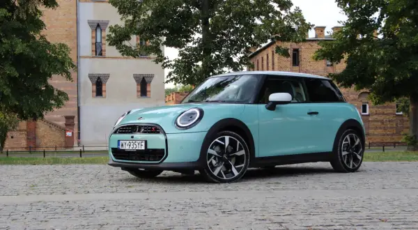 Turkusowy Mini Cooper stoi na brukowanej drodze przed ceglanym budynkiem.